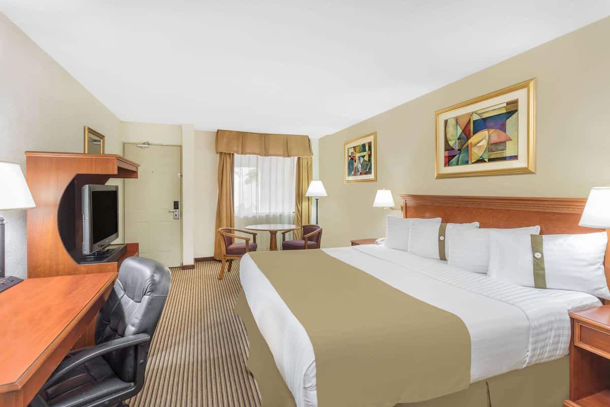 A hotel room with a bed, a desk with a television, two chairs, a table, and a painting on the wall.