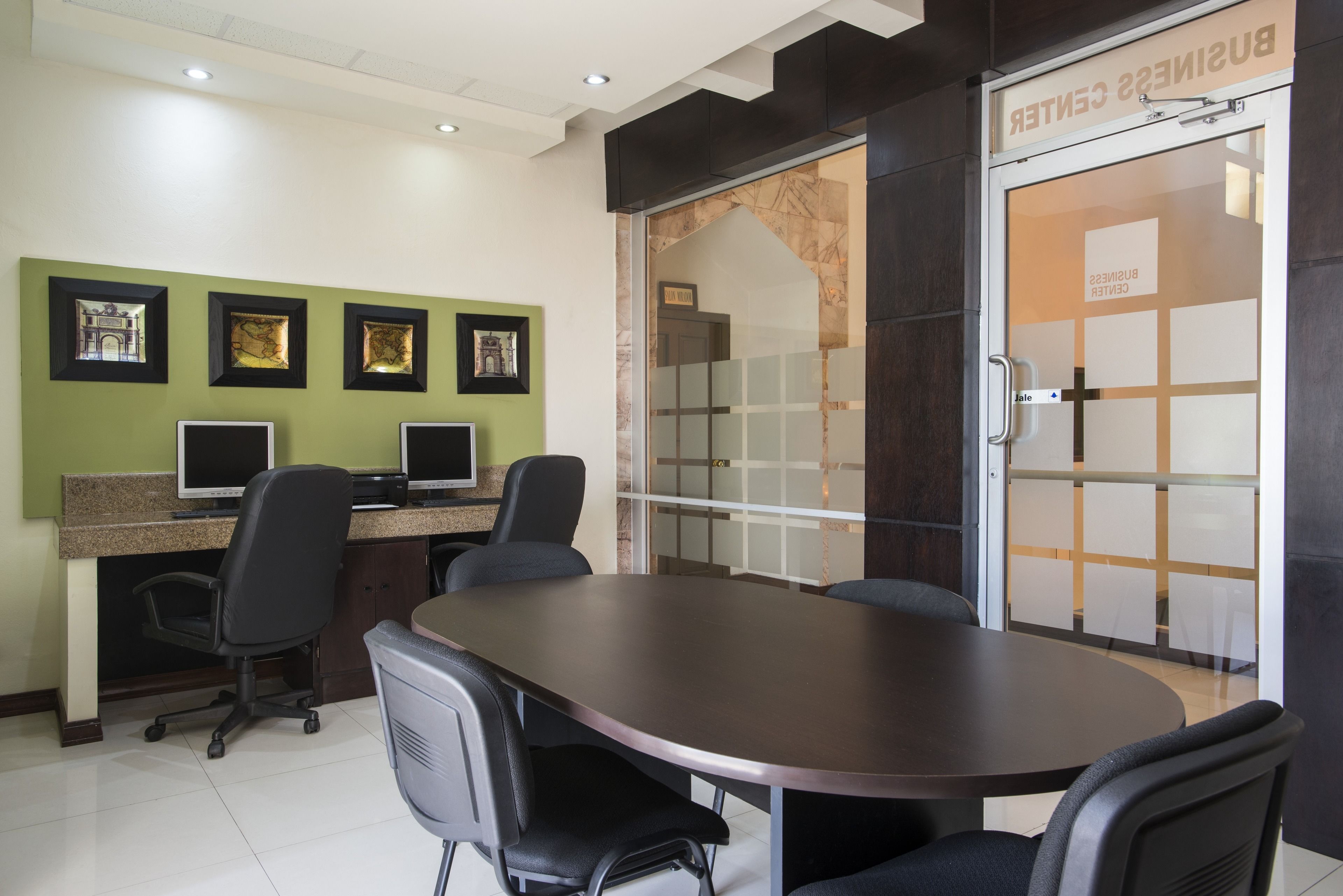 Een vergaderruimte met een grote ovale tafel, vier stoelen, twee computermonitoren en ingelijste foto's aan de muur.