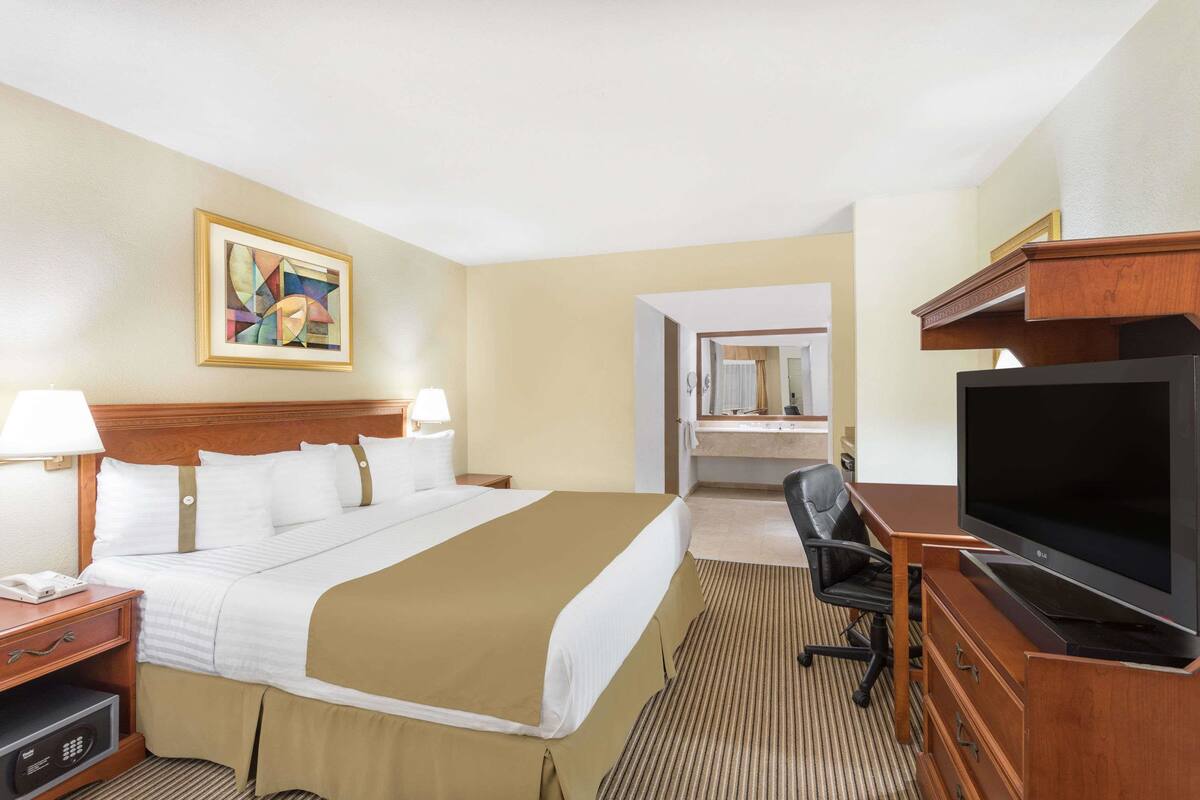 A hotel room with a large bed, bedside tables, a desk with a TV, a chair, and a bathroom visible through an open door.