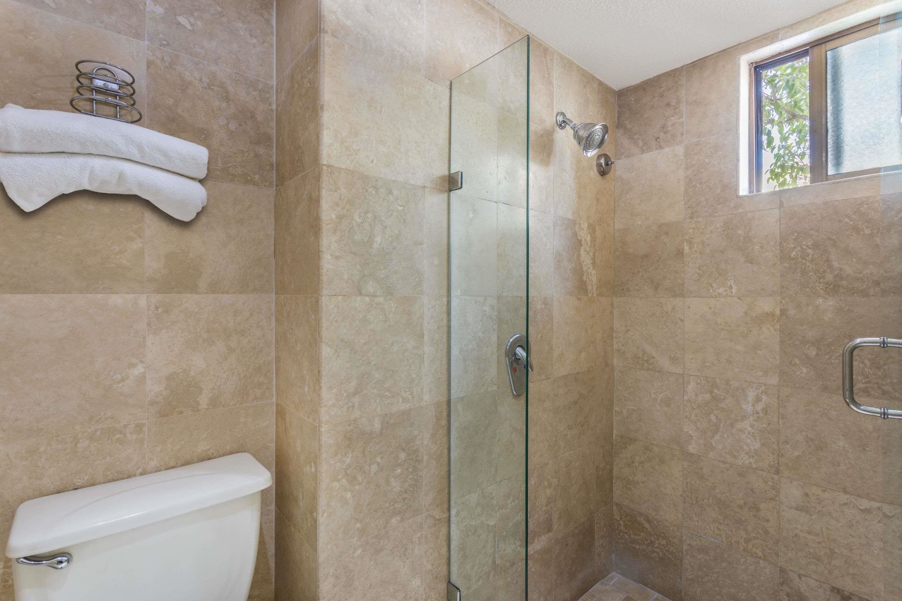 A bathroom with a glass-enclosed shower, beige tiles, a toilet, a towel rack, and a window.