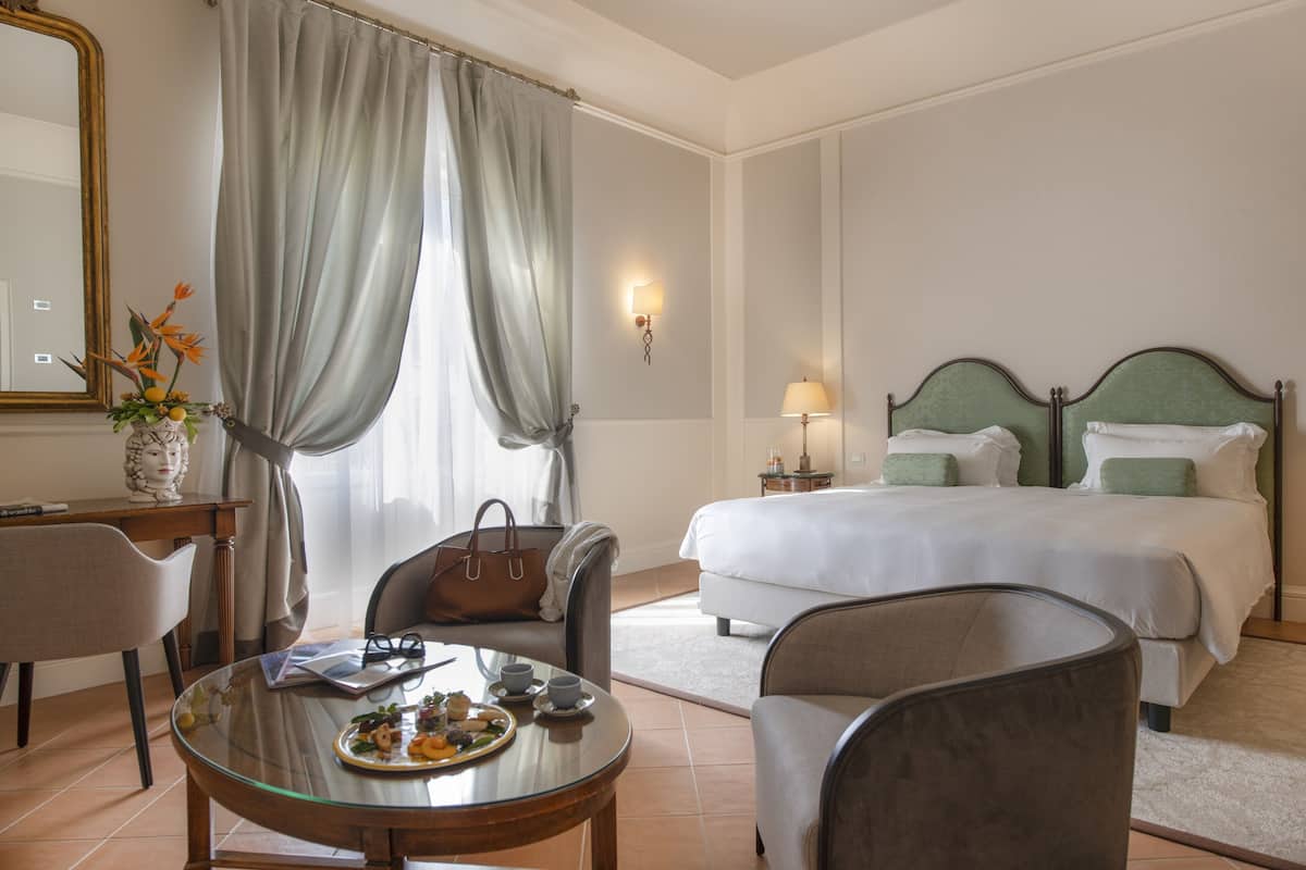 Habitación de hotel con cama, dos sillas, una mesa con un plato de comida, un florero con flores y una lámpara.