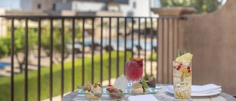 Una mesa con varios aperitivos y una bebida, frente a un balcón con vista a edificios y follaje.
