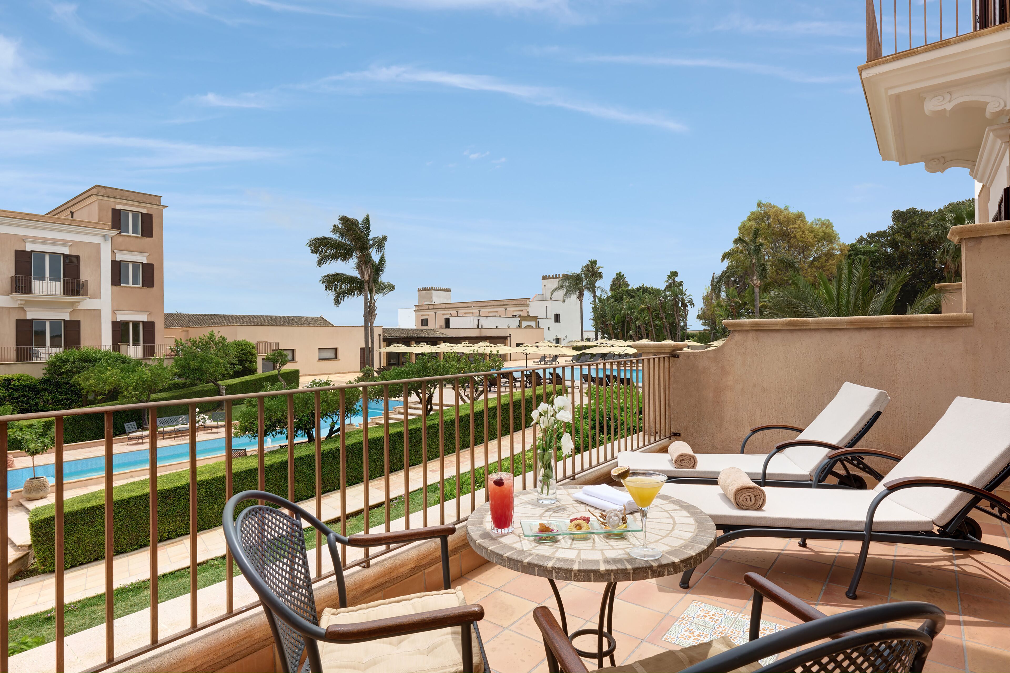 A balcony with lounge chairs, a table with drinks, a pool, and a view of a building and greenery.
