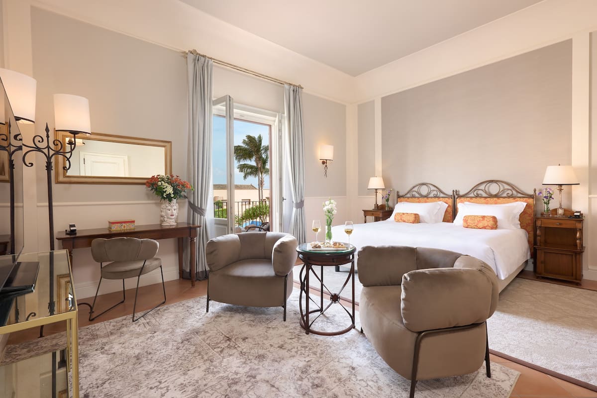 Una habitación de hotel con cama, dos sillones, una mesita, un escritorio y un ventanal grande con cortinas.