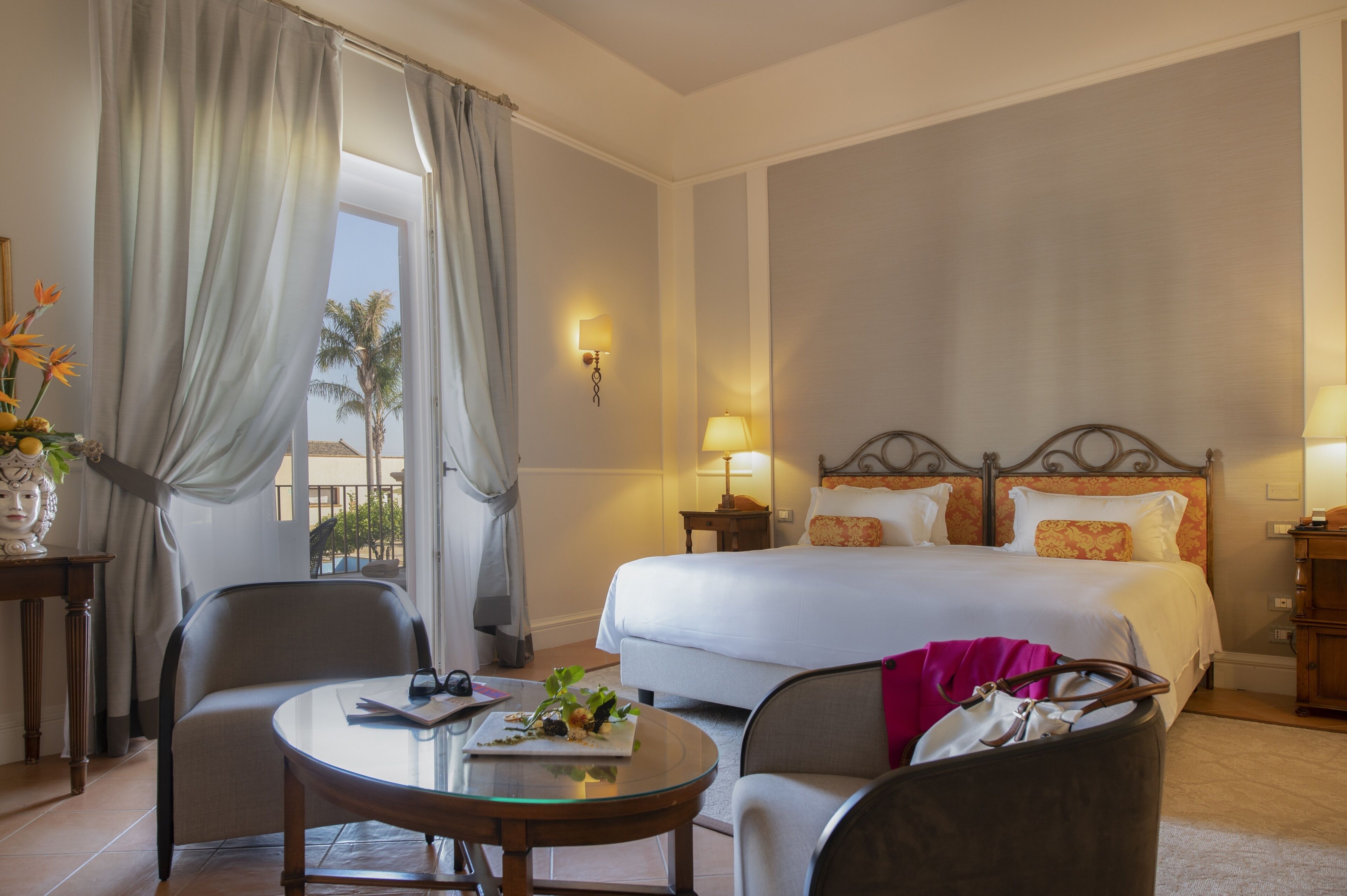 Habitación de hotel con una cama, dos mesitas de noche con lámparas, una mesa redonda de vidrio, una silla y vistas a palmeras a través de la ventana.