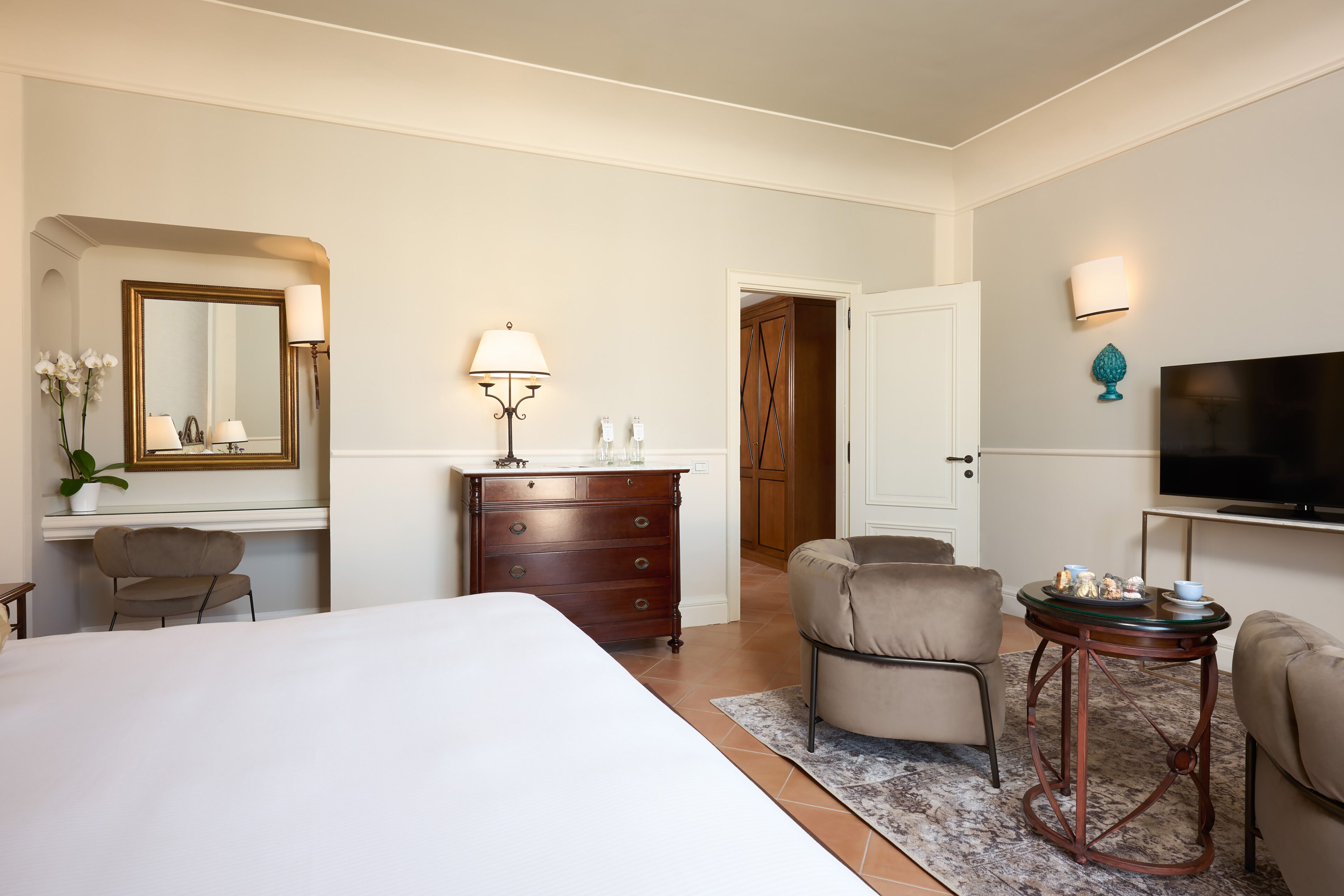 Una camera d'albergo con un letto grande, una cassettiera, una scrivania con lampada, una sedia, un tavolino con la televisione e una porta che dà su un'altra stanza.