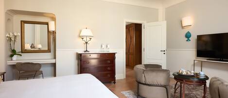 Una camera d'albergo con un letto grande, una cassettiera, una scrivania con lampada, una sedia, un tavolino con la televisione e una porta che dà su un'altra stanza.
