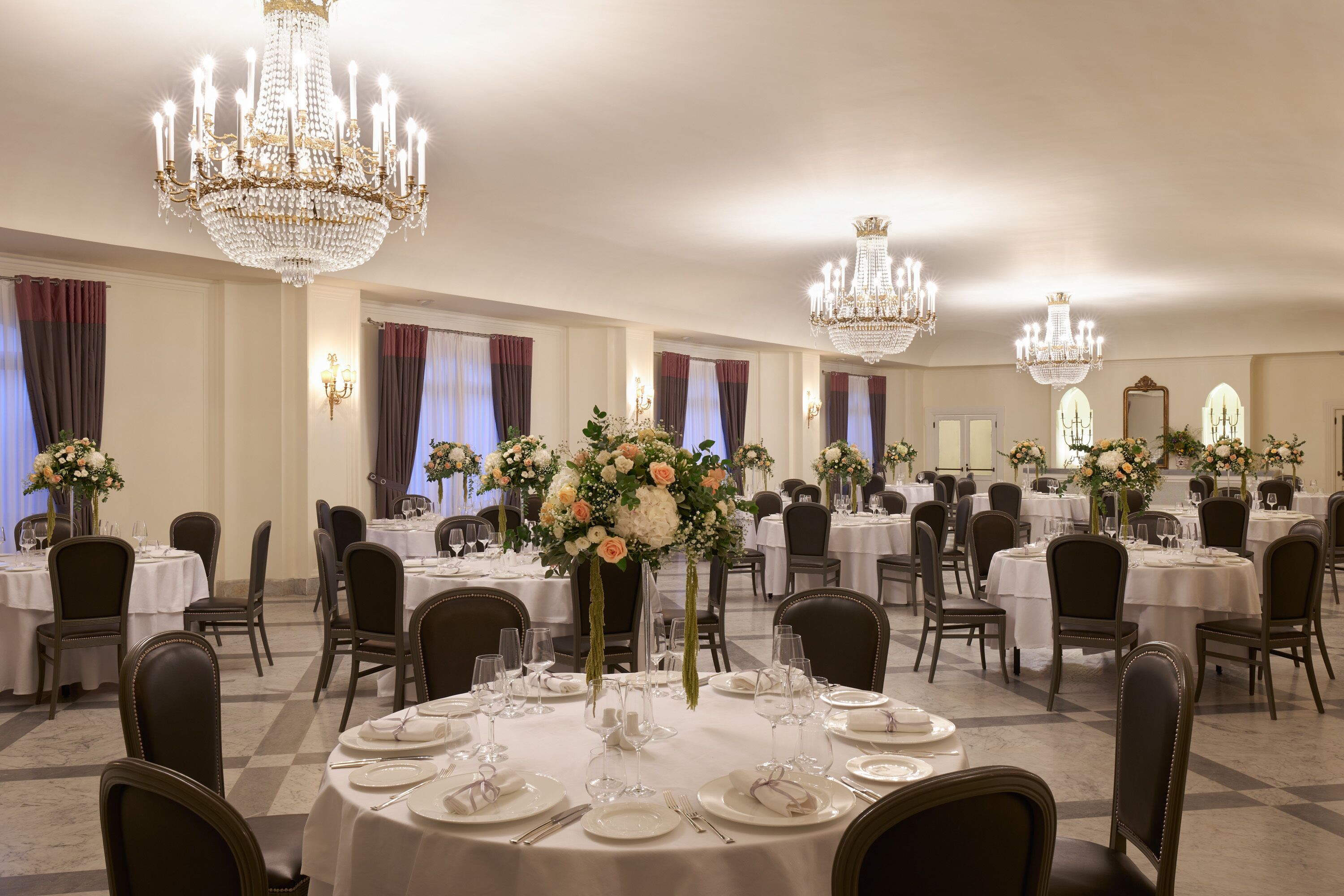 A banquet hall with round tables, chairs, chandeliers, and floral centerpieces.