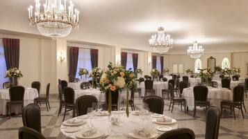 A banquet hall with round tables, chairs, chandeliers, and floral centerpieces.