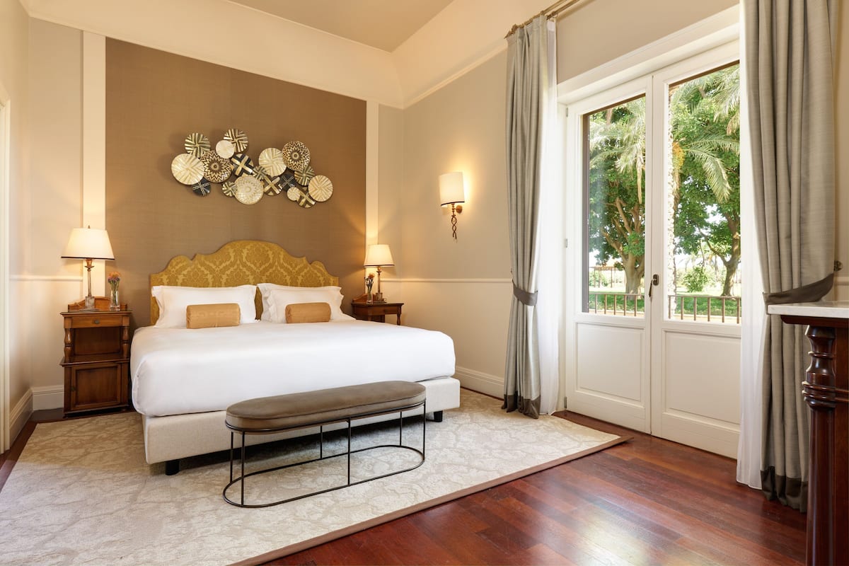 Un dormitorio con una cama grande, mesitas de noche con lámparas, un adorno en la pared y vistas a palmeras a través de la puerta.