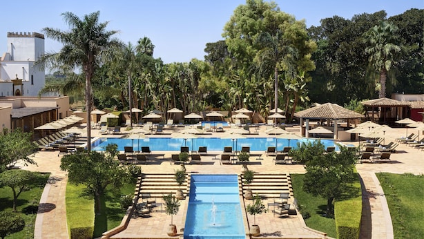 A resort pool area with a central water feature, surrounded by lounge chairs and umbrellas, and palm trees. - Almar Giardino di Costanza Mazara del Vallo Resort & Spa (Mazara del Vallo)