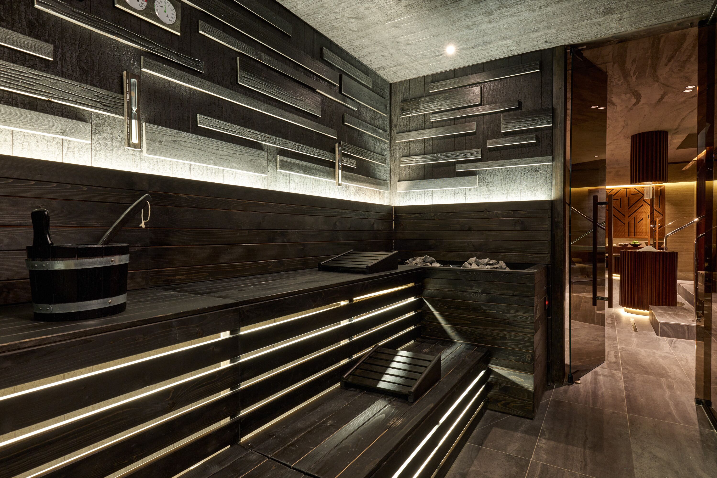 A modern sauna with wooden benches and a stone floor, featuring a wall-mounted clock and a small heater on the bench.