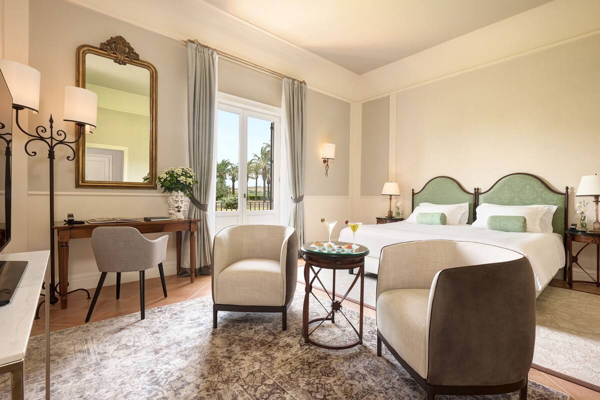 Habitación de hotel con cama, dos sillones, escritorio, espejo y vista a palmeras.