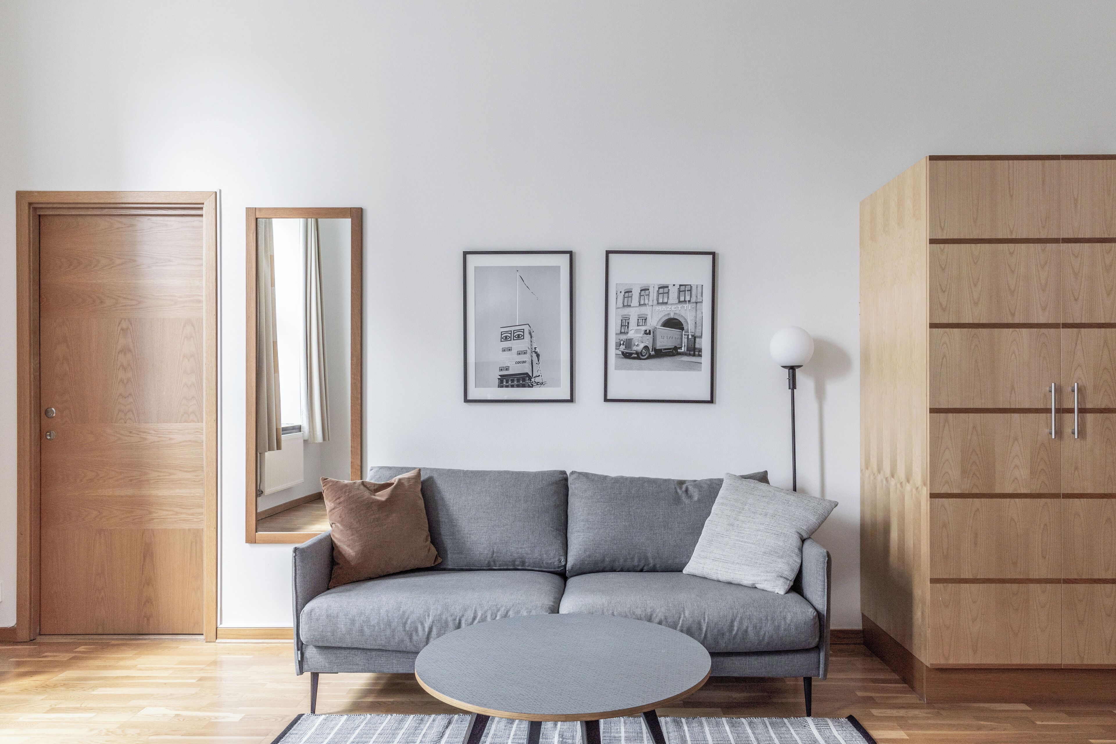 Ett modernt vardagsrum med en grå soffa, ett runt soffbord och en trägarderob. På väggen hänger två inramade tavlor och en spegel.