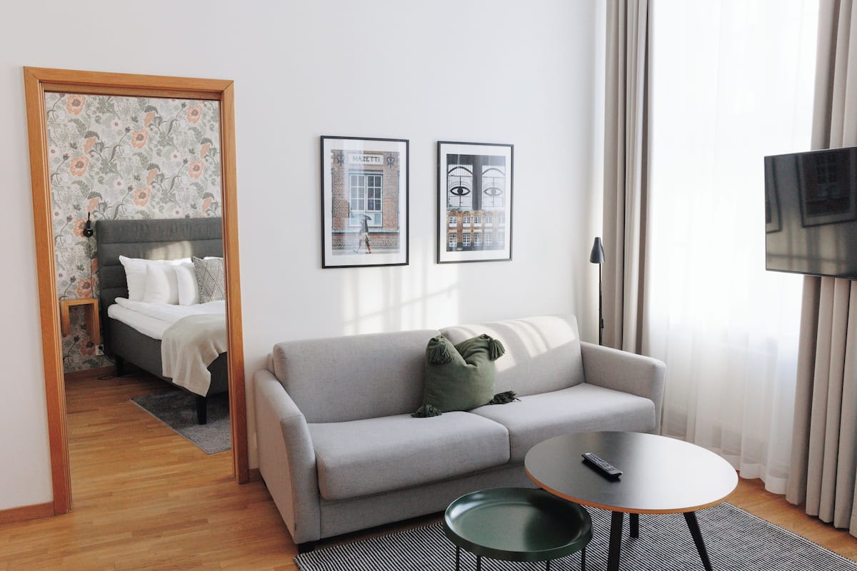 Moderni hotellihuone, jossa on harmaa sohva, pieni pyöreä pöytä ja harmaalla päätyosalla varustettu sänky. Seinällä on kaksi kehystettyä kuvaa, oikealla puolella on televisio ja ikkunassa on verhot.
