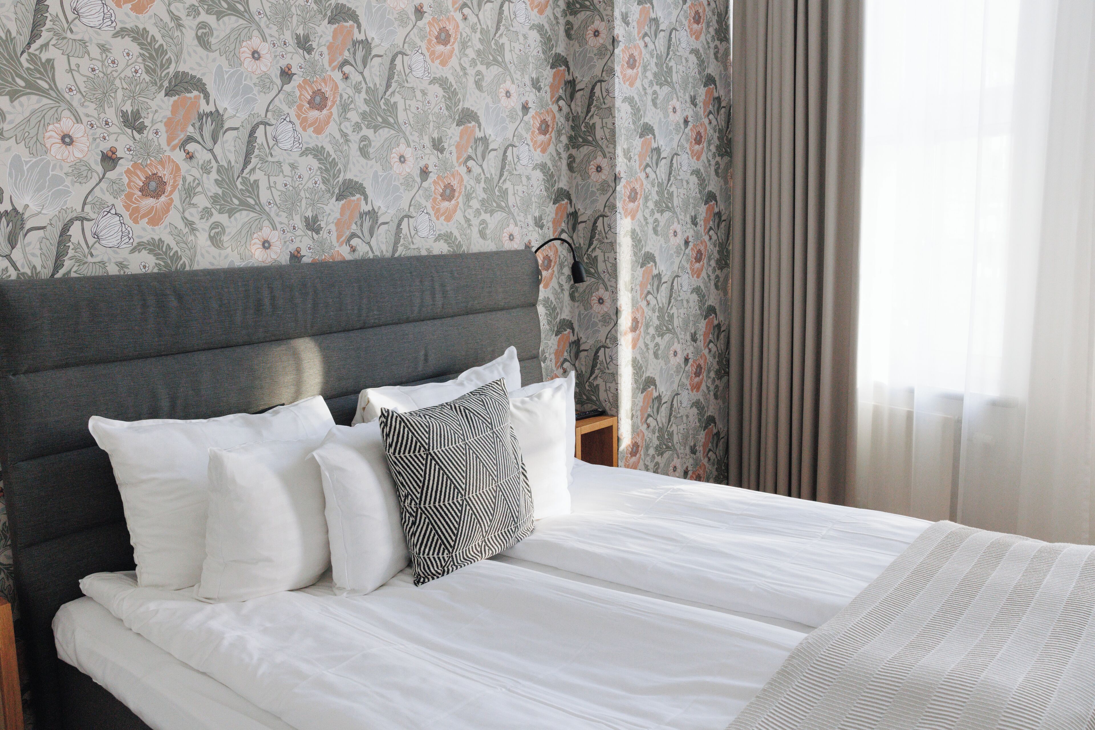 A neatly made bed with white linens and a grey headboard, set against a floral wallpaper.