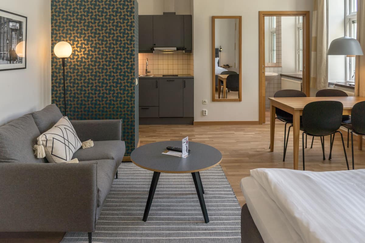 Moderni hotellihuone, jossa on harmaa sohva, pyöreä sohvapöytä, sänky, ruokapöytä tuoleineen, keittiöalue ja suuri peili.