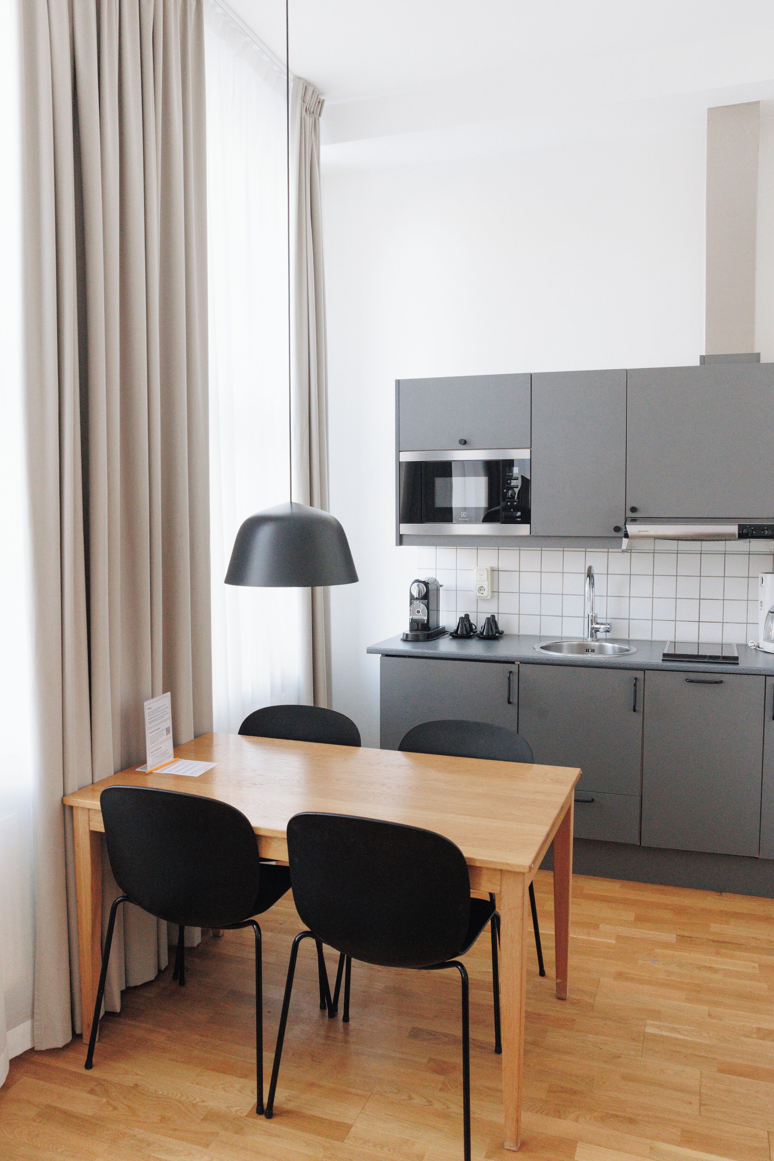 A modern kitchen with grey cabinets, a wooden dining table, two black chairs, a black pendant lamp, and a white tiled backsplash.