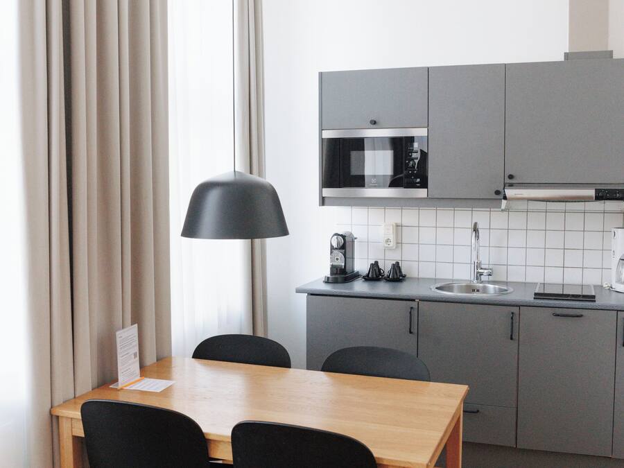 A modern kitchen with grey cabinets, a wooden dining table, two black chairs, a black pendant lamp, and a white tiled backsplash.