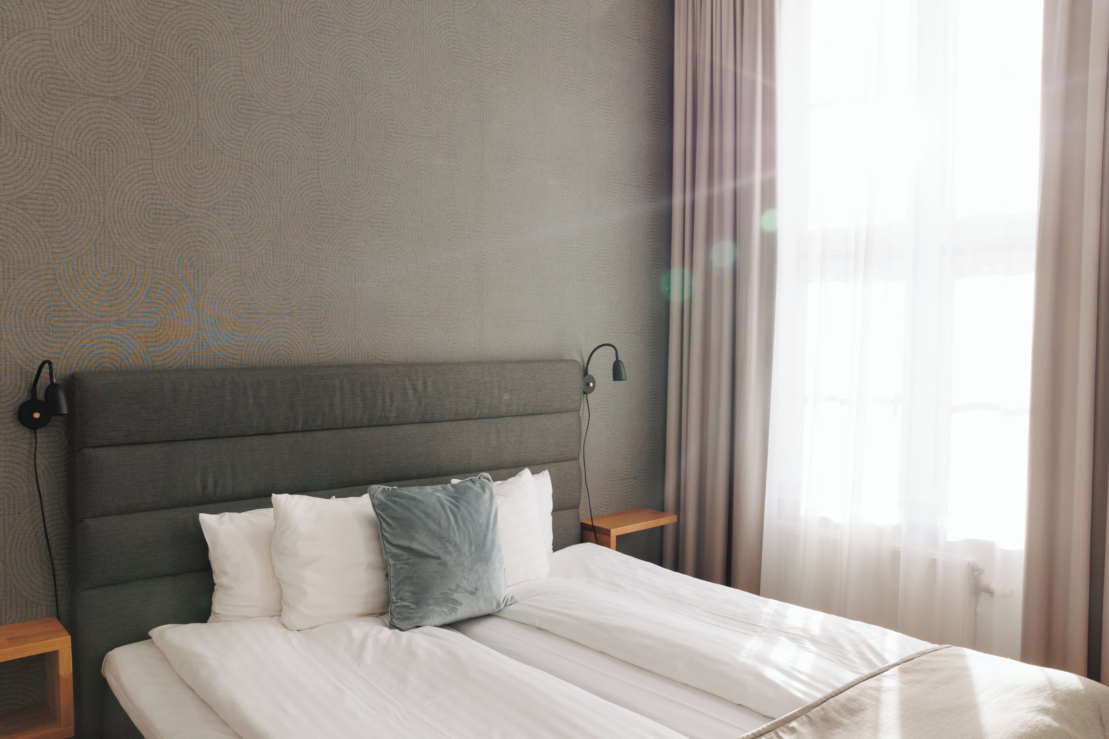 A neatly made bed with white linens and a grey headboard, two bedside tables with lamps, and a window with sheer curtains.