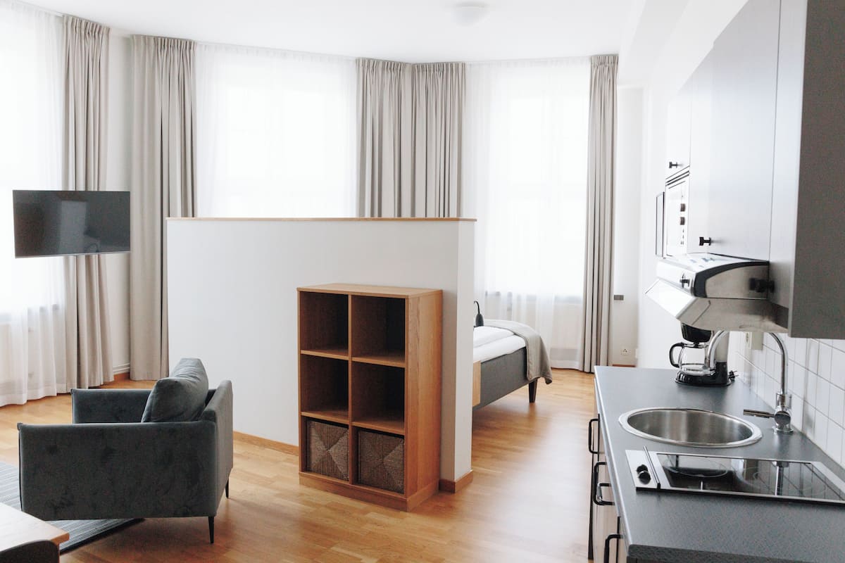 Moderni hotellihuone, jossa on sohva, televisio, sänky, pieni keittiö ja suuri ikkuna, jossa on verhot.