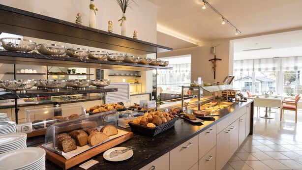 A buffet area with various food items, a seating area with a table and chairs, and a view of the outside through large windows. - Huberhof (Allershausen)