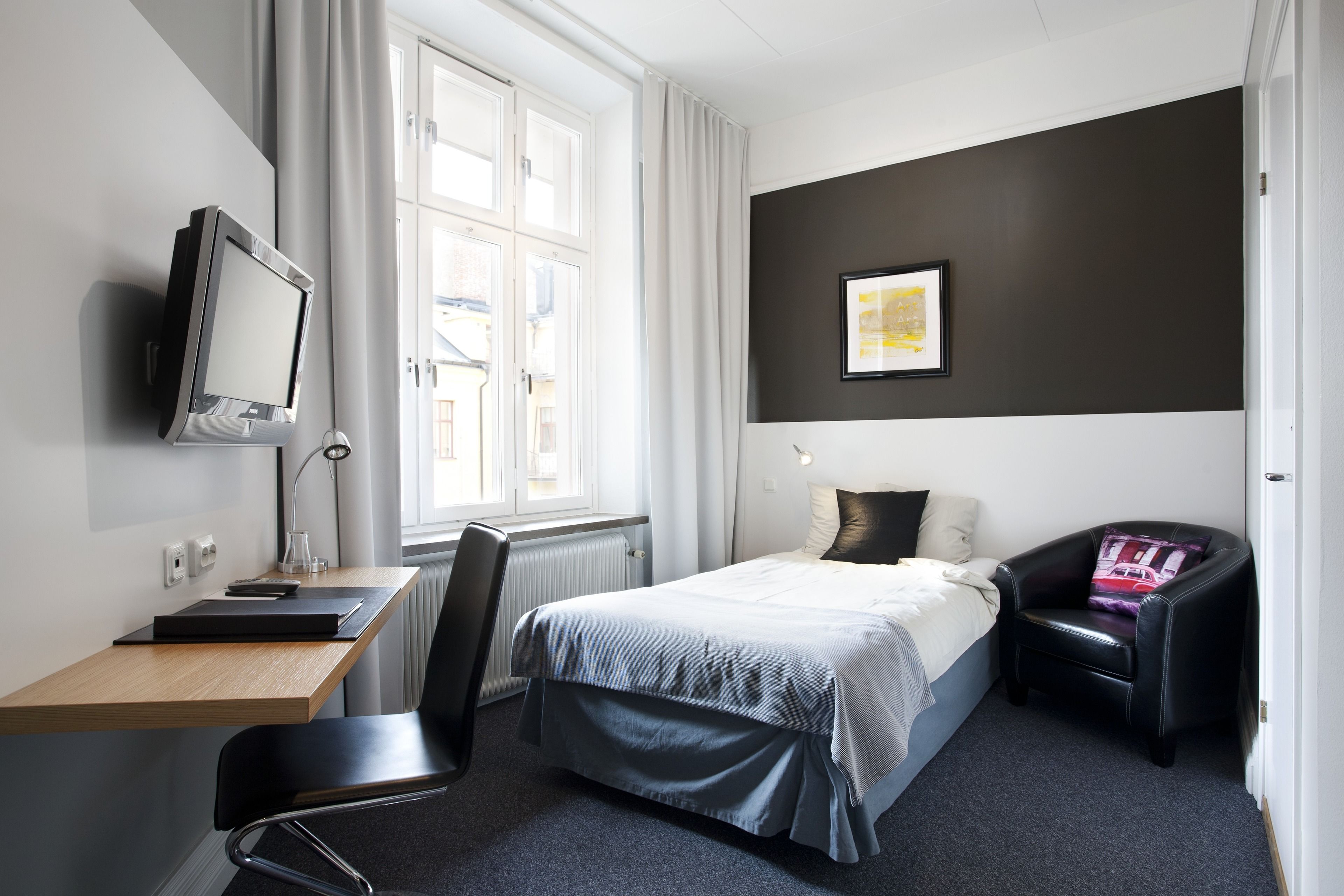 Ett hotellrum med en säng, ett skrivbord med en dator, en stol, en tv och ett fönster med gardiner.