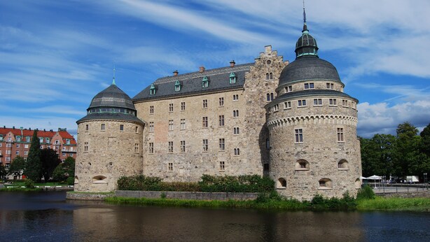 A historic stone castle with two prominent towers and a large central building, situated by a calm body of water.