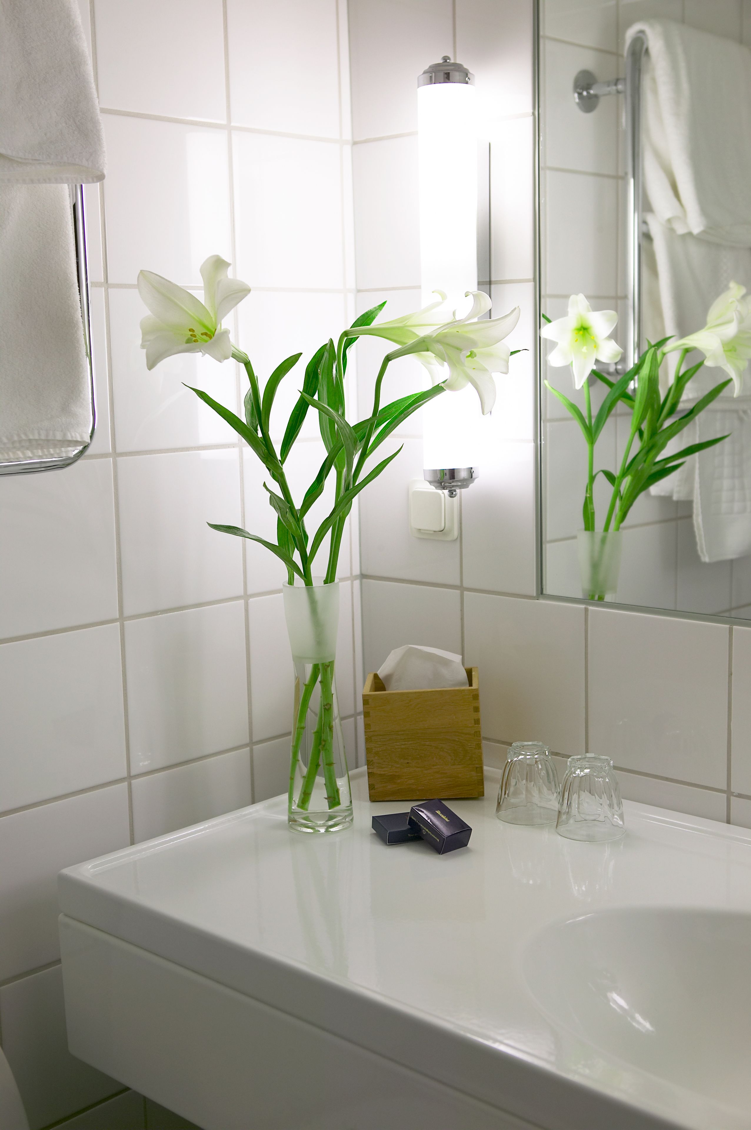 Un baño con un lavamanos blanco, un espejo y un florero con flores blancas.