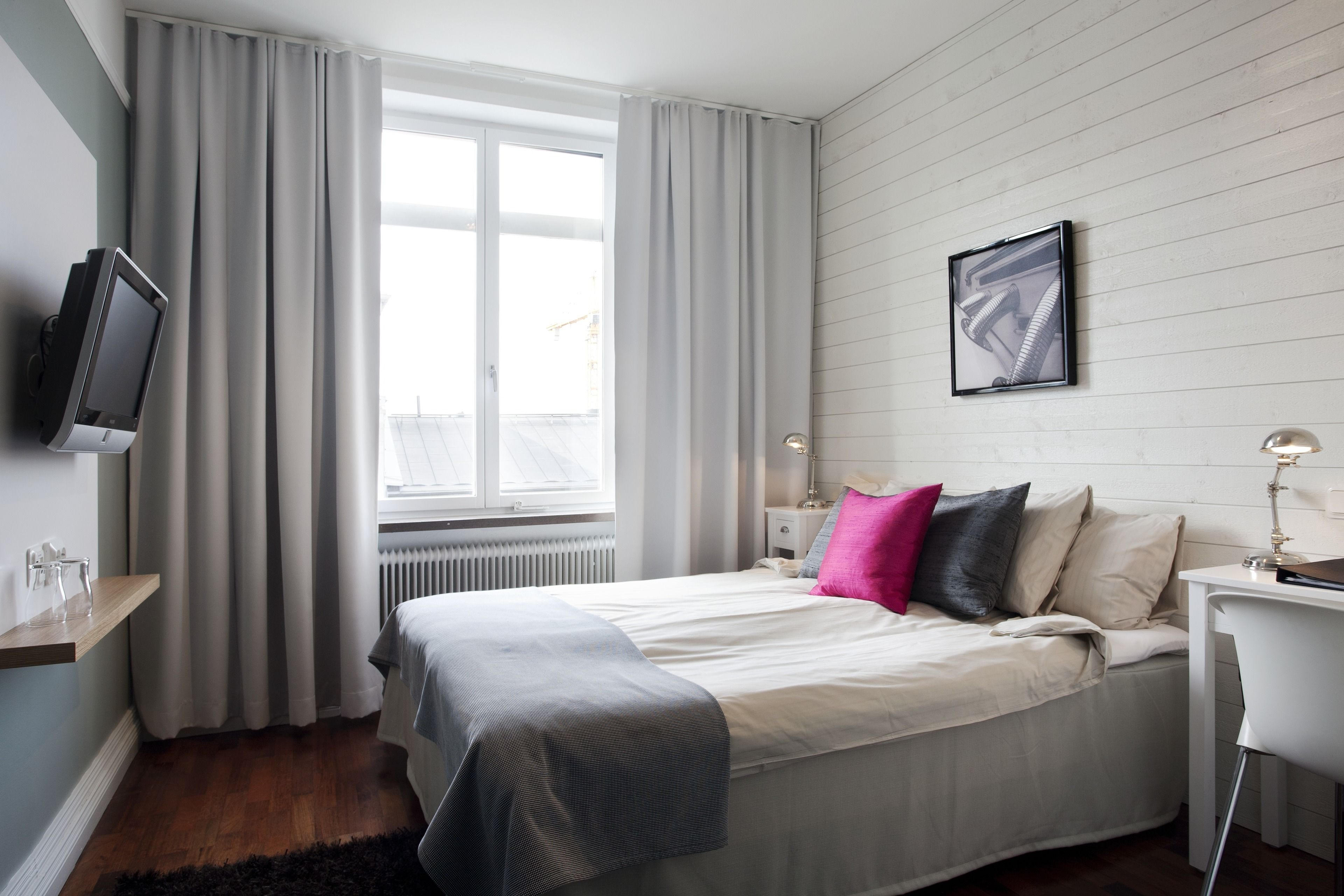 Un dormitorio con cama, televisor montado en la pared, escritorio con silla y ventana con cortinas.