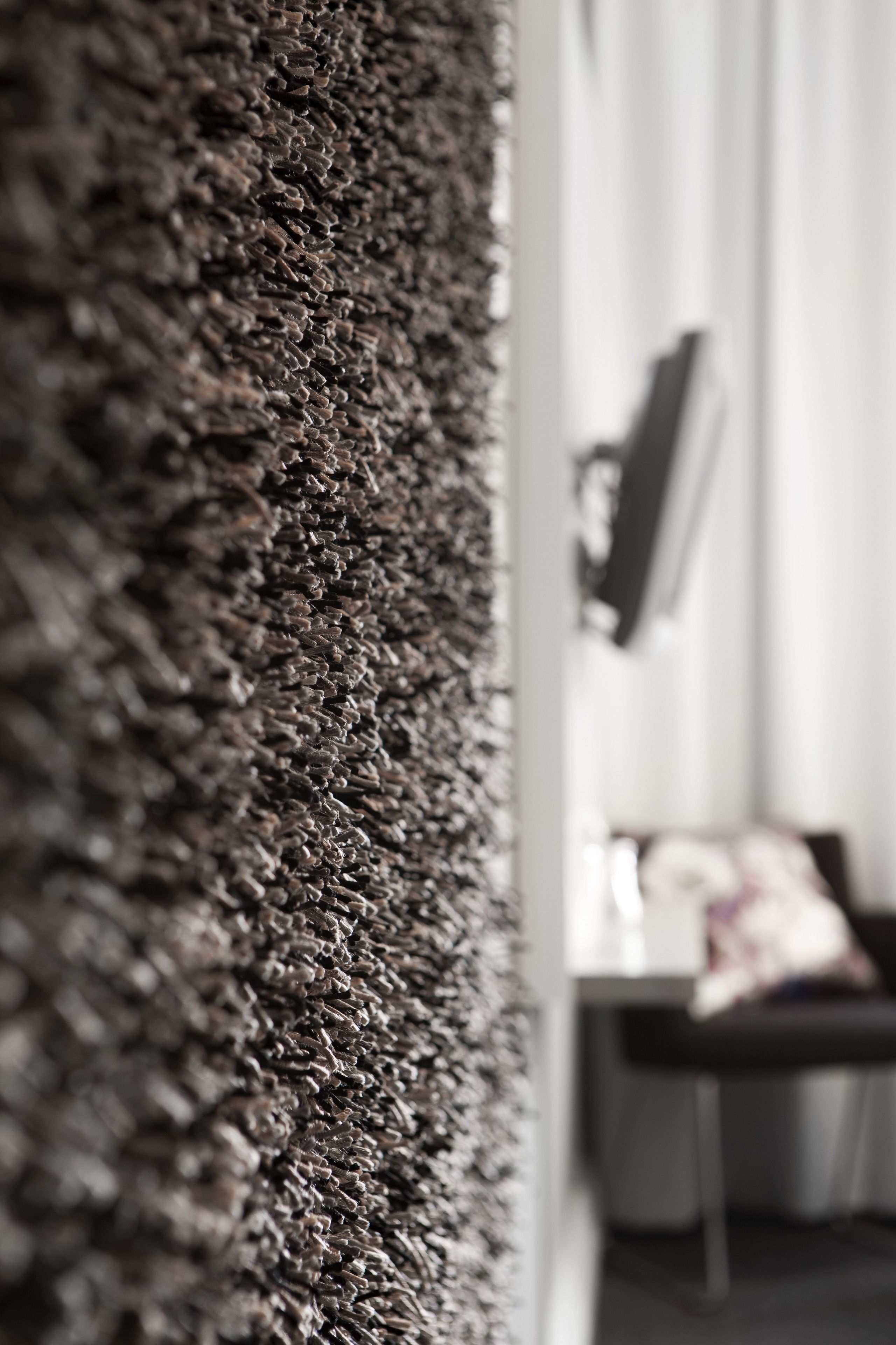 A close-up of a textured wall, likely a textured carpet or wall covering, with a blurred background suggesting a room with a chair and a table.