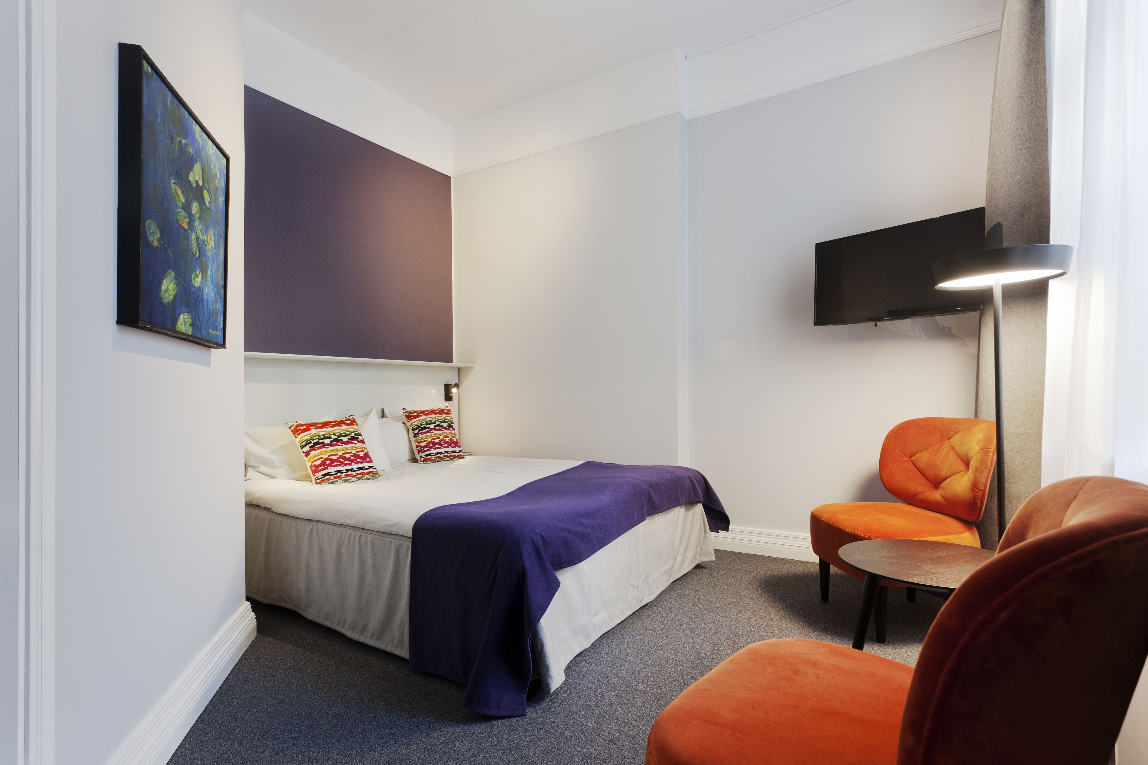 Un dormitorio con una cama, una silla, un televisor montado en la pared y un cuadro.