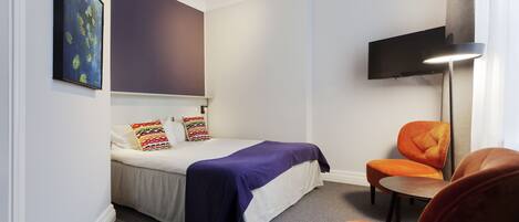 Un dormitorio con una cama, una silla, un televisor montado en la pared y un cuadro.