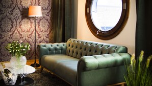 A room with a green sofa, a round window, a patterned wallpaper, a floor lamp, and a small table with a vase and glasses.