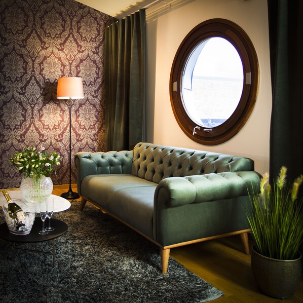 A room with a green sofa, a round window, a patterned wallpaper, a floor lamp, and a small table with a vase and glasses.