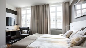 A modern hotel room with a large bed, a desk with a flat-screen TV, a chair, and large windows with curtains.
