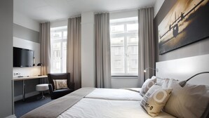 A hotel room with two beds, a desk, a chair, a TV, and large windows with curtains.