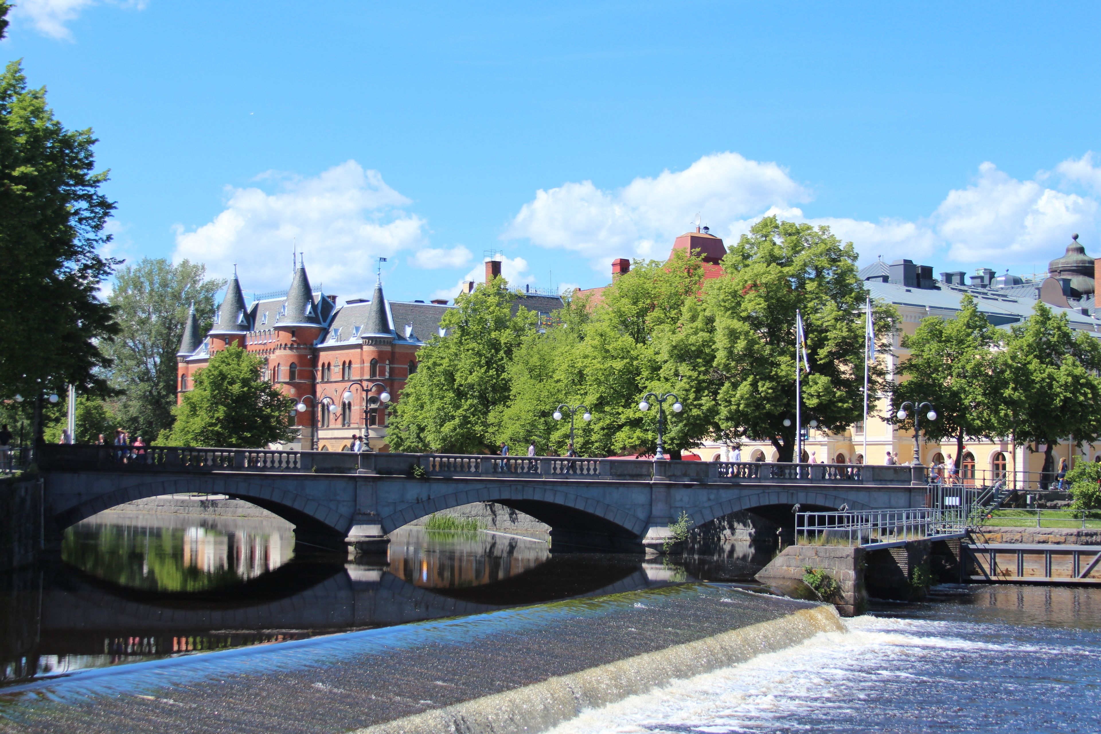 En stenbro över en flod, där byggnader och träd speglas i vattnet.