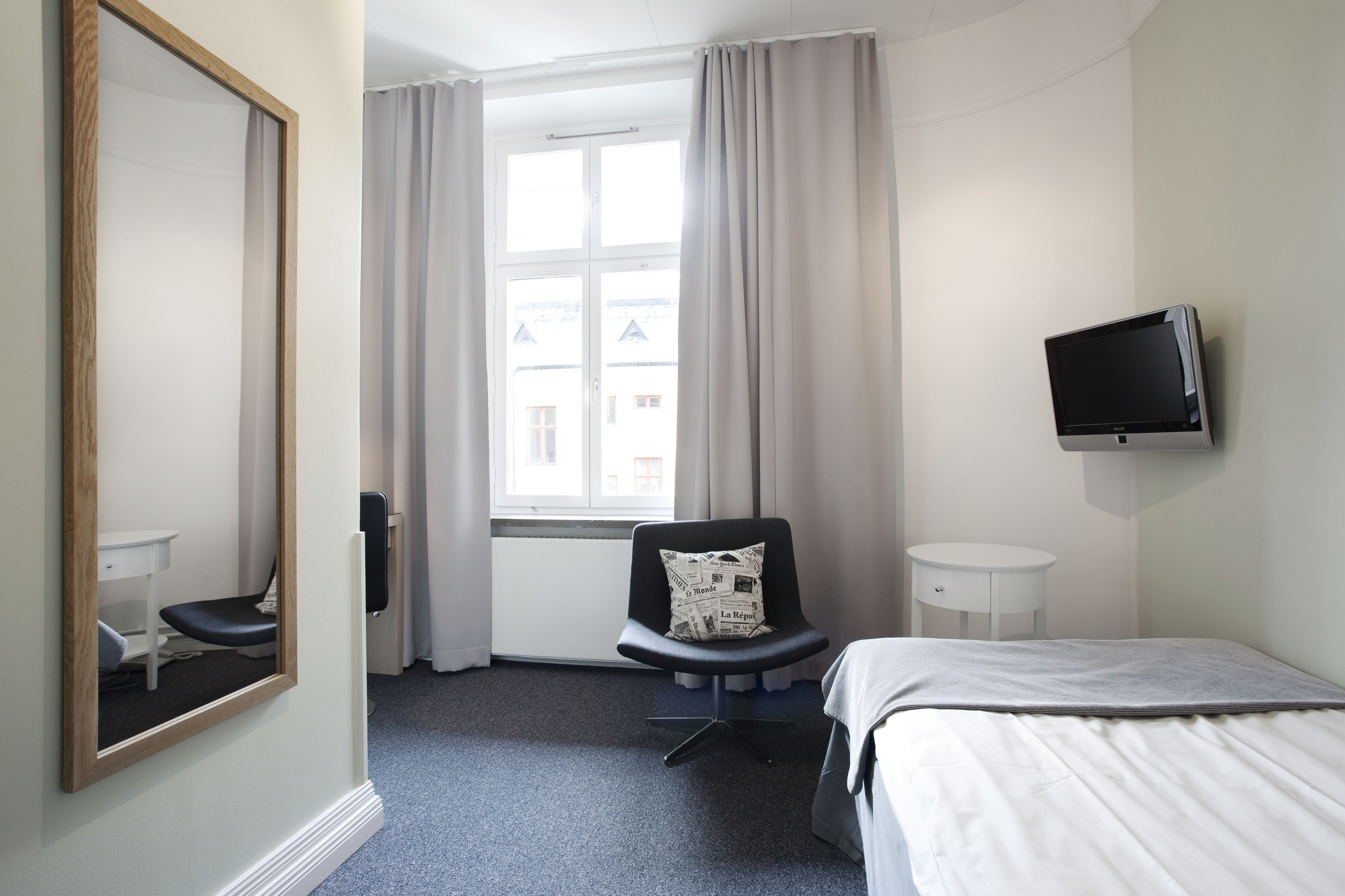 Habitación de hotel con cama, una silla con un cojín con periódico, una mesita, un televisor montado en la pared y una ventana con cortinas.