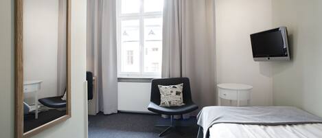 Habitación de hotel con cama, una silla con un cojín con periódico, una mesita, un televisor montado en la pared y una ventana con cortinas.