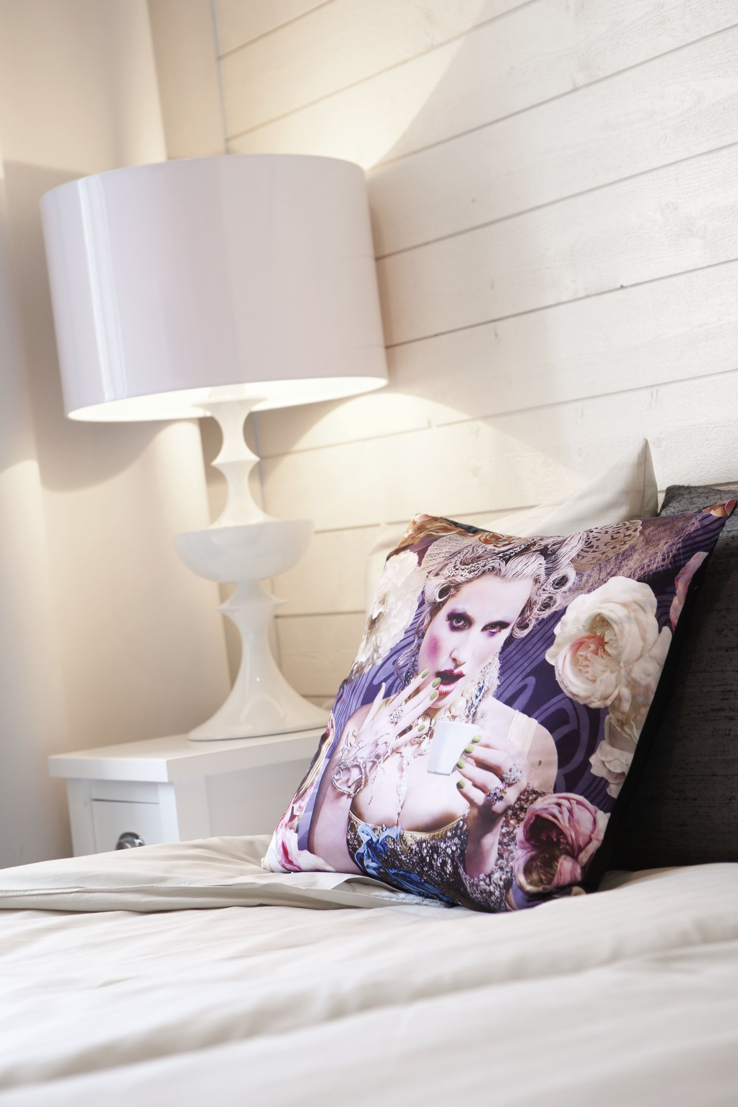 Un lit avec un coussin décoratif, une table de chevet blanche et une lampe blanche.