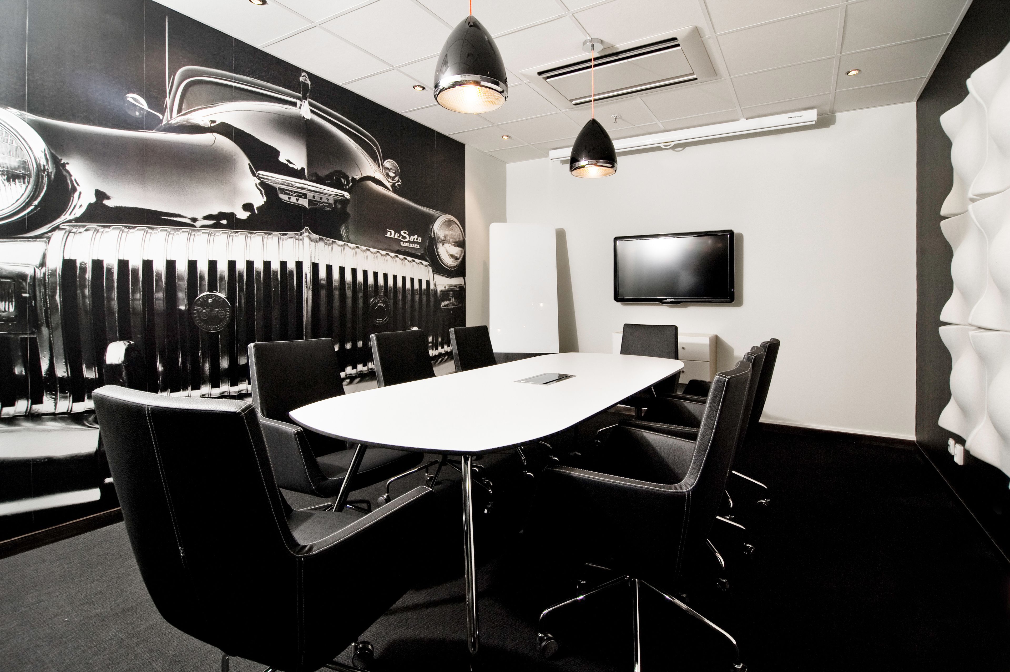 A conference room with a white table, black chairs, a mounted TV, and a large wall mural of a car.