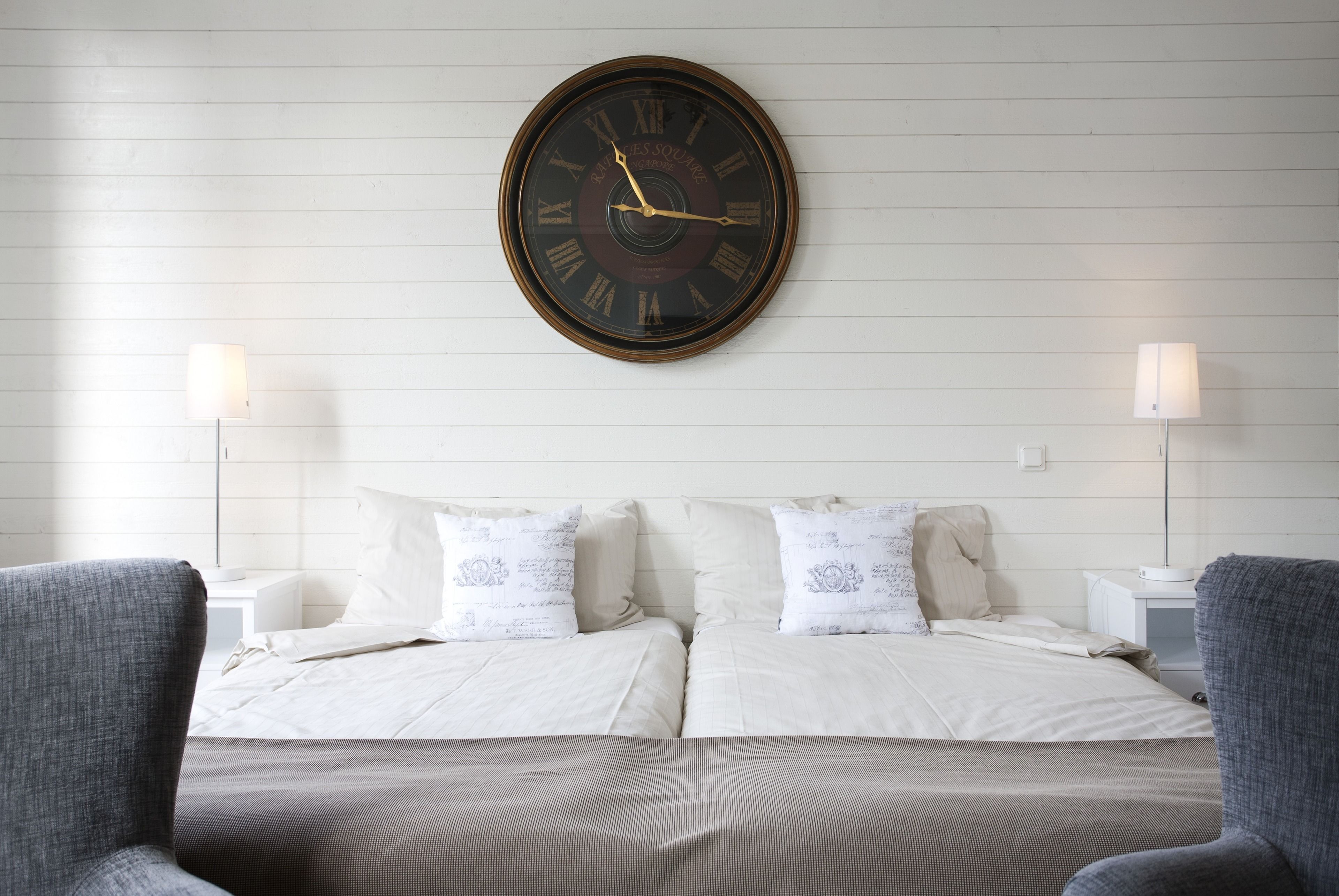 A hotel room with a large wall clock, two bedside tables with lamps, a bed with white bedding, and two gray armchairs.