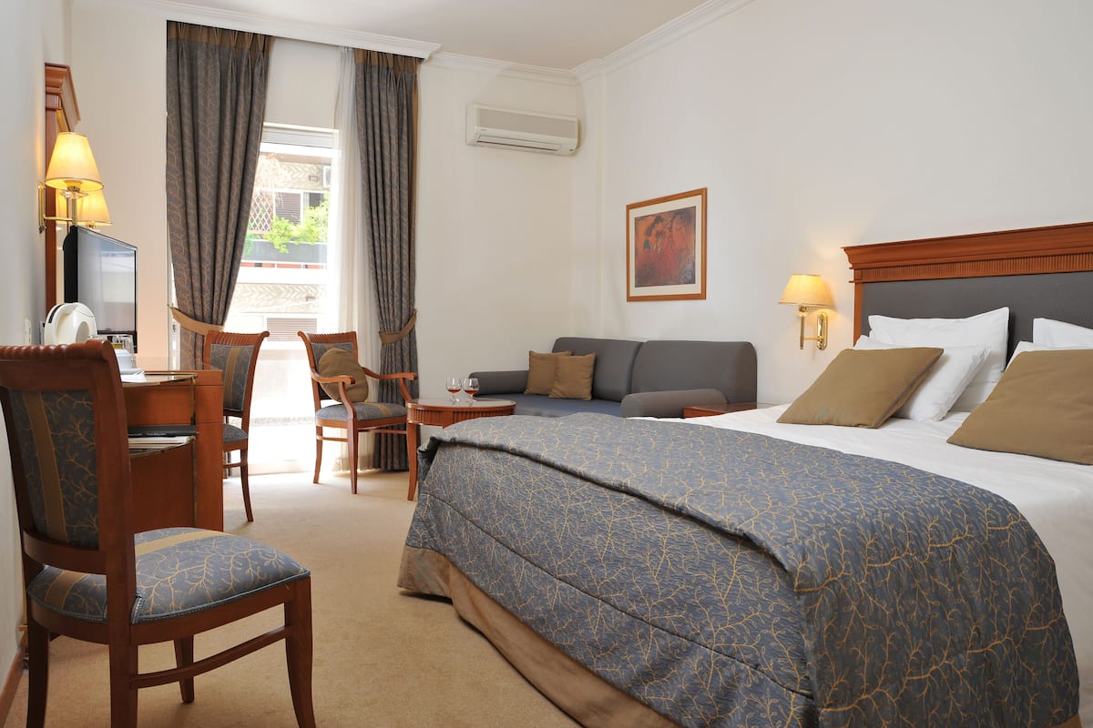 A hotel room with a large bed, a desk with a computer, a chair, a sofa, and a window with curtains.