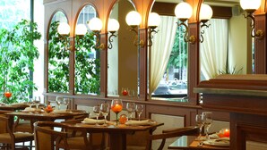 A restaurant with multiple tables, each set with wine glasses and plates, and a large window with curtains and greenery visible outside. - Athens Atrium Hotel and Suites (Athens)
