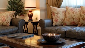 A cozy living room with a leather sofa, floral cushions, a small table with a book and a candle, and a decorative plant. - Athens Atrium Hotel and Suites (Athens)