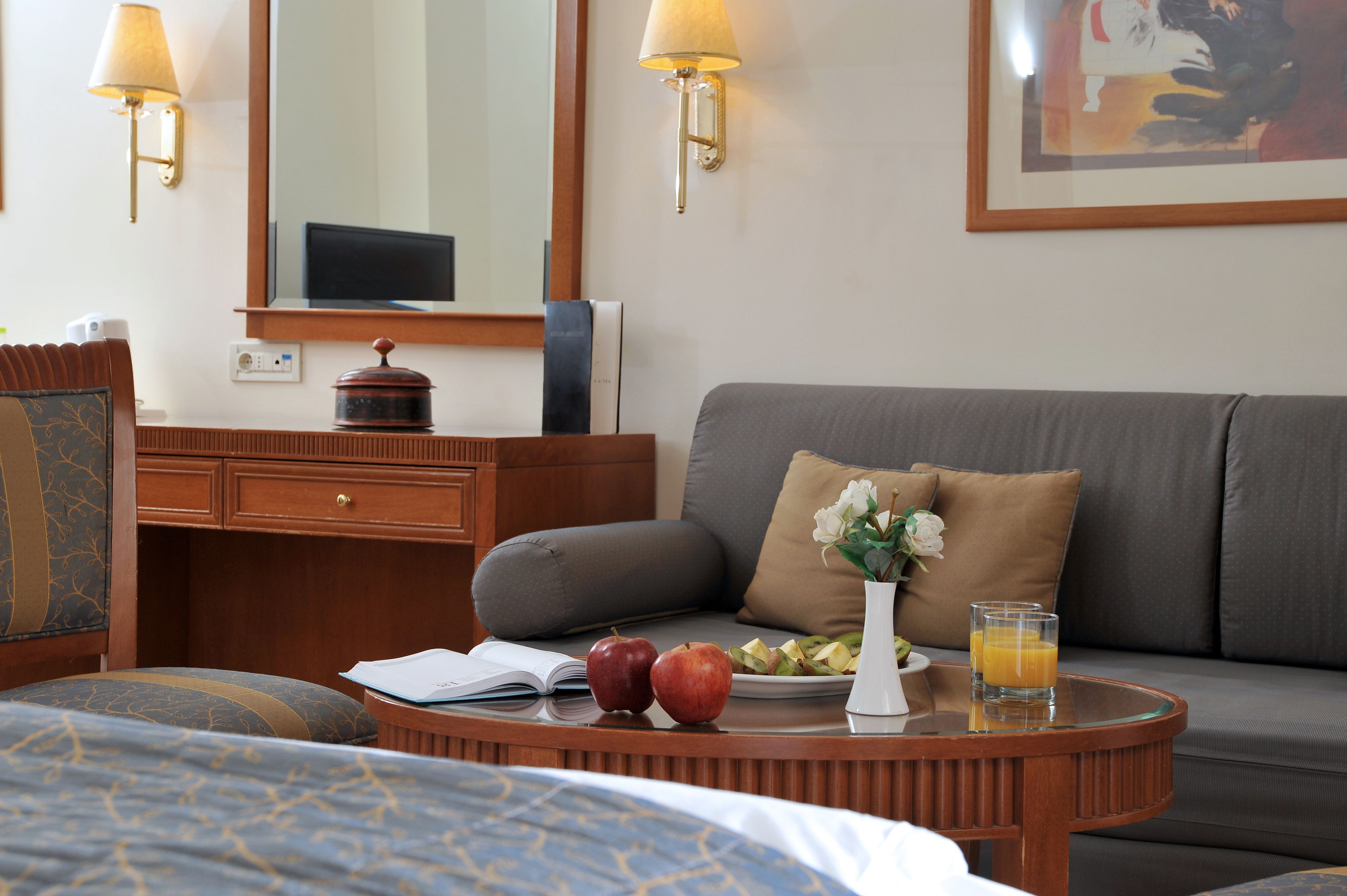 A hotel room with a bed, a round wooden table with breakfast items, a grey sofa, a wooden desk, and a mirror.