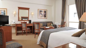 A hotel room with a bed, desk, chairs, a television, and a large window with curtains. - Athens Atrium Hotel and Suites (Athens)