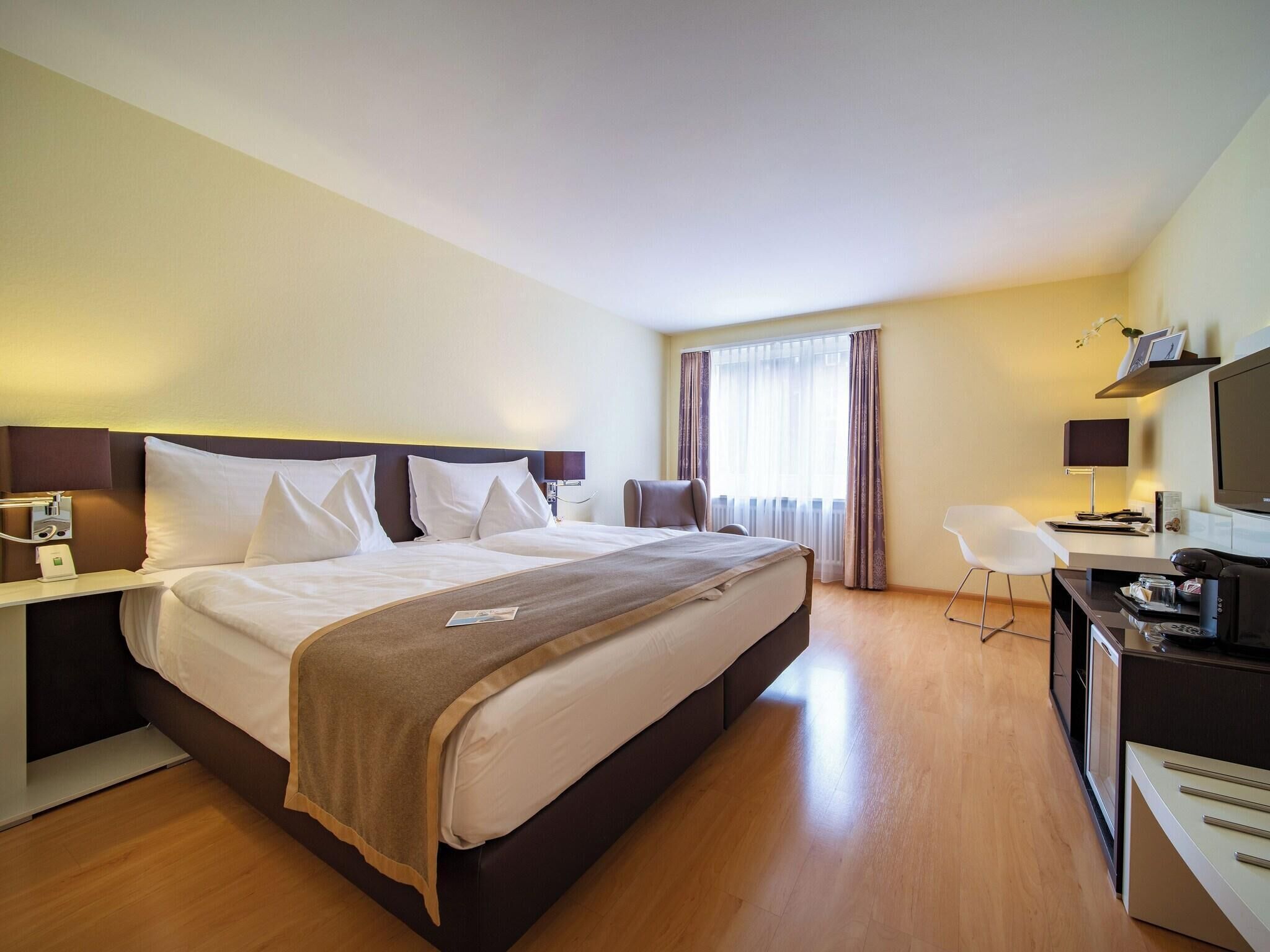 Une chambre d’hôtel avec un grand lit, des tables de chevet, un bureau avec une chaise, une télévision et une fenêtre avec des rideaux.