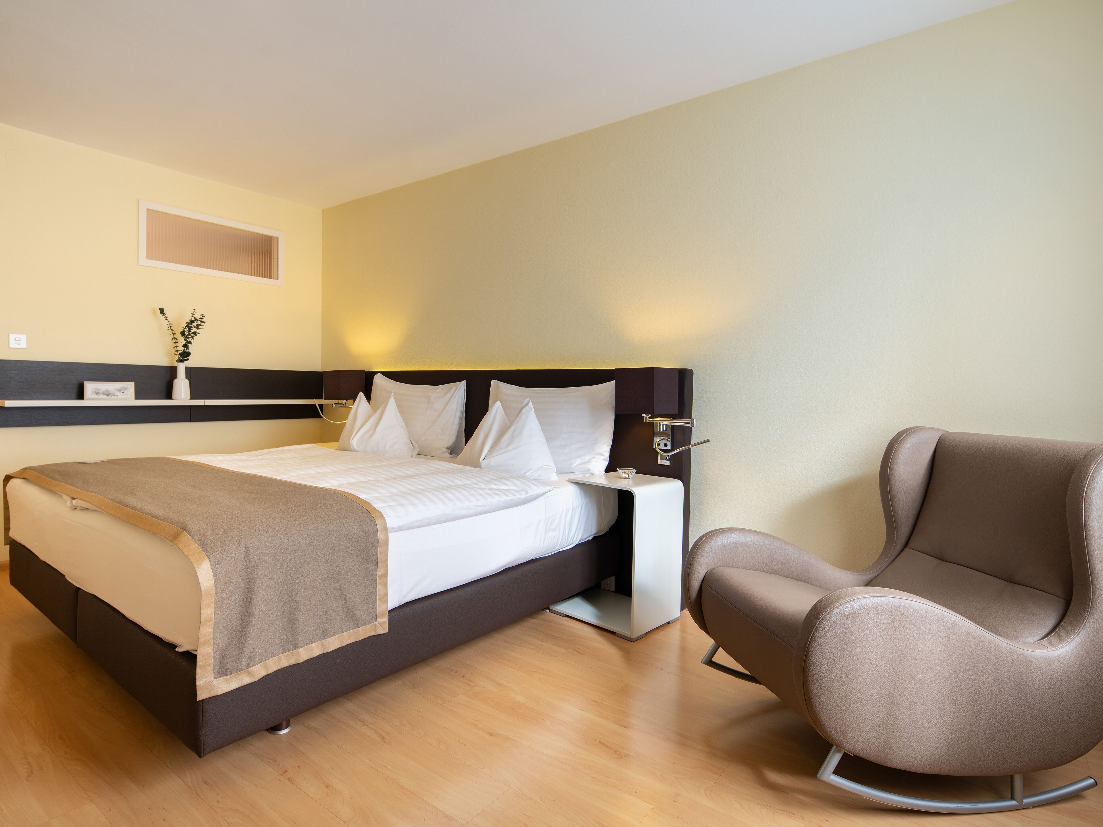 A hotel room with a bed, bedside table, chair, and a wall-mounted shelf with a plant.